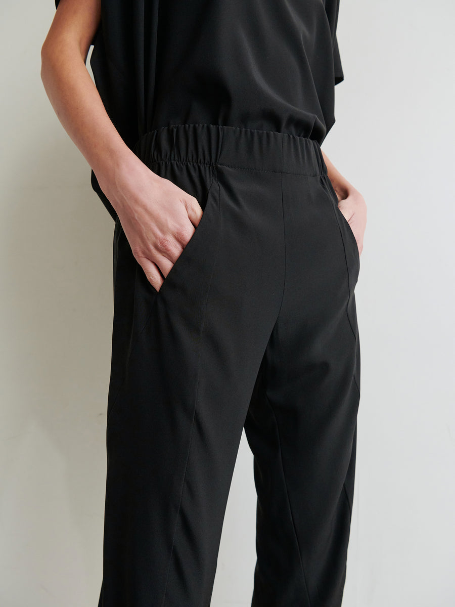 A person stands with hands in the pockets of loose-fitting Cropped Akeo Pants from Zero + Maria Cornejo, made from stretch silk charmeuse in black, featuring an elastic waistband. They are wearing a matching black top. Only the lower torso and upper legs are visible against a light background.