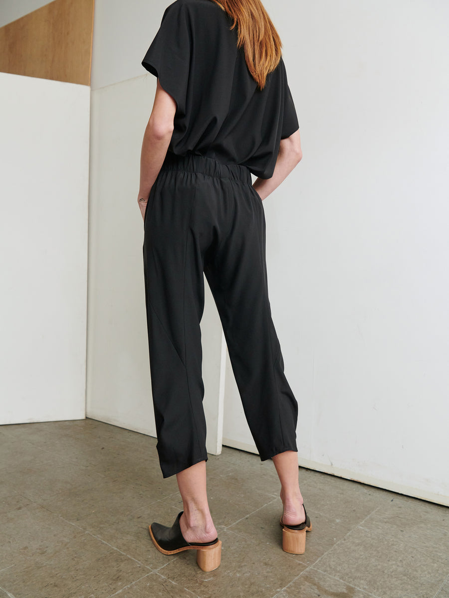 A person with long hair stands facing away from the camera, wearing a loose-fitting black outfit with cropped pants and short sleeves. The Cropped Akeo Pant by Zero + Maria Cornejo features an elastic waist for added comfort. They are also wearing heeled sandals. The background is a minimal interior space with light-colored walls and a tiled floor.