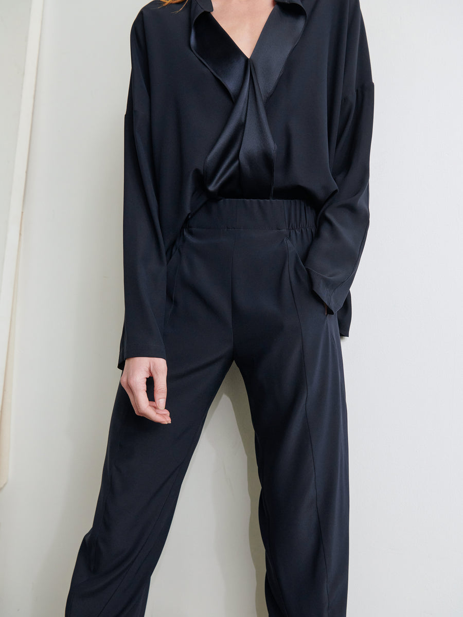 A person wearing a stylish, loose-fitting black satin blouse tucked into high-waisted Cropped Akeo Pants by Zero + Maria Cornejo, posing with one hand in their pocket against a plain background.