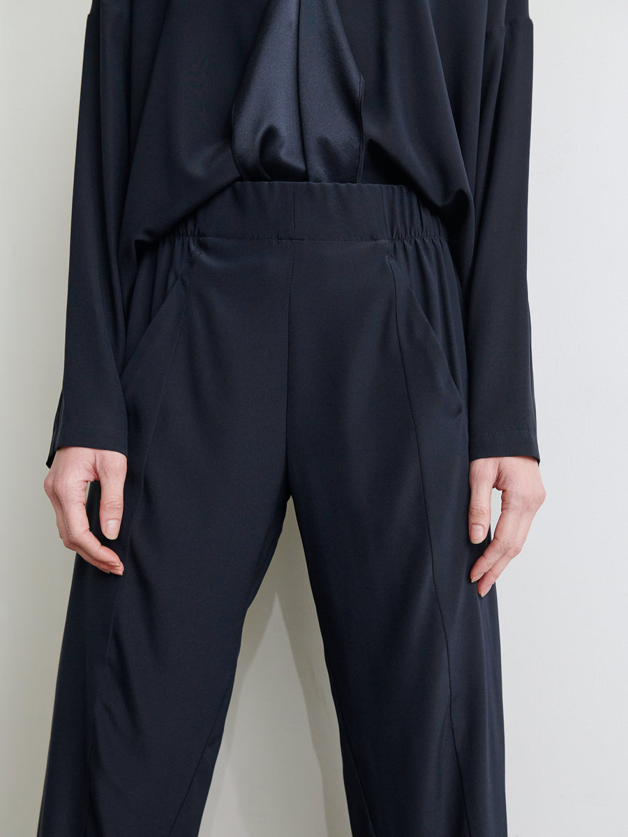 A person wearing the Cropped Akeo Pant from Zero + Maria Cornejo, which are loose-fitting and dark-colored with an elastic waist, along with a partially visible matching top is shown in this image. Their hands are relaxed at their sides, and the background is plain and neutral.