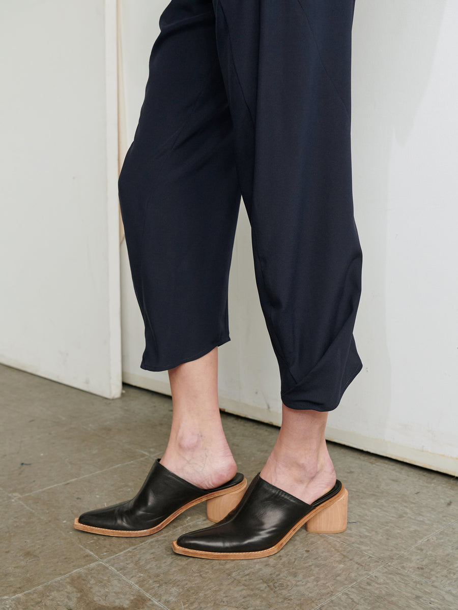 A person wearing the Cropped Akeo Pant from Zero + Maria Cornejo with an elastic waist and black mules with wooden block heels stands on a tiled floor next to a white wall.