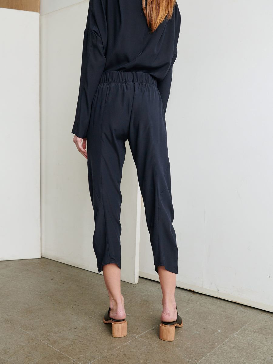 A person is facing a corner of a room, standing with their back to the camera. They are wearing a black top and the Cropped Akeo Pant from Zero + Maria Cornejo, which features an elastic waist and is made from stretch silk charmeuse. Their outfit is completed with wooden block-heeled sandals. The room has a minimalist style with white walls and a gray floor.