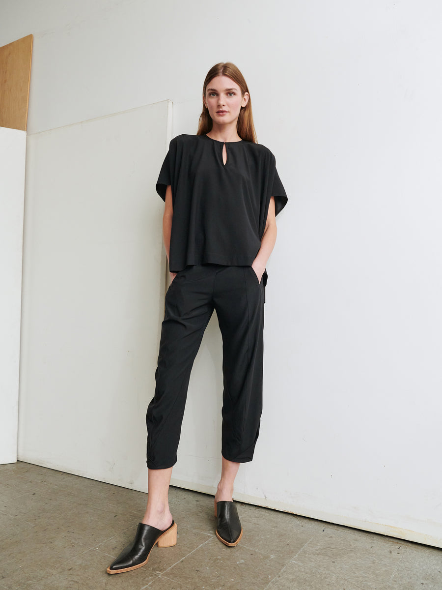 A person with long hair stands against a white wall, wearing the Rae Poncho Top by Zero + Maria Cornejo paired with black cropped pants. They are also wearing black, backless slip-on shoes with wooden heels. The floor is concrete, and a wooden panel is propped against the wall.