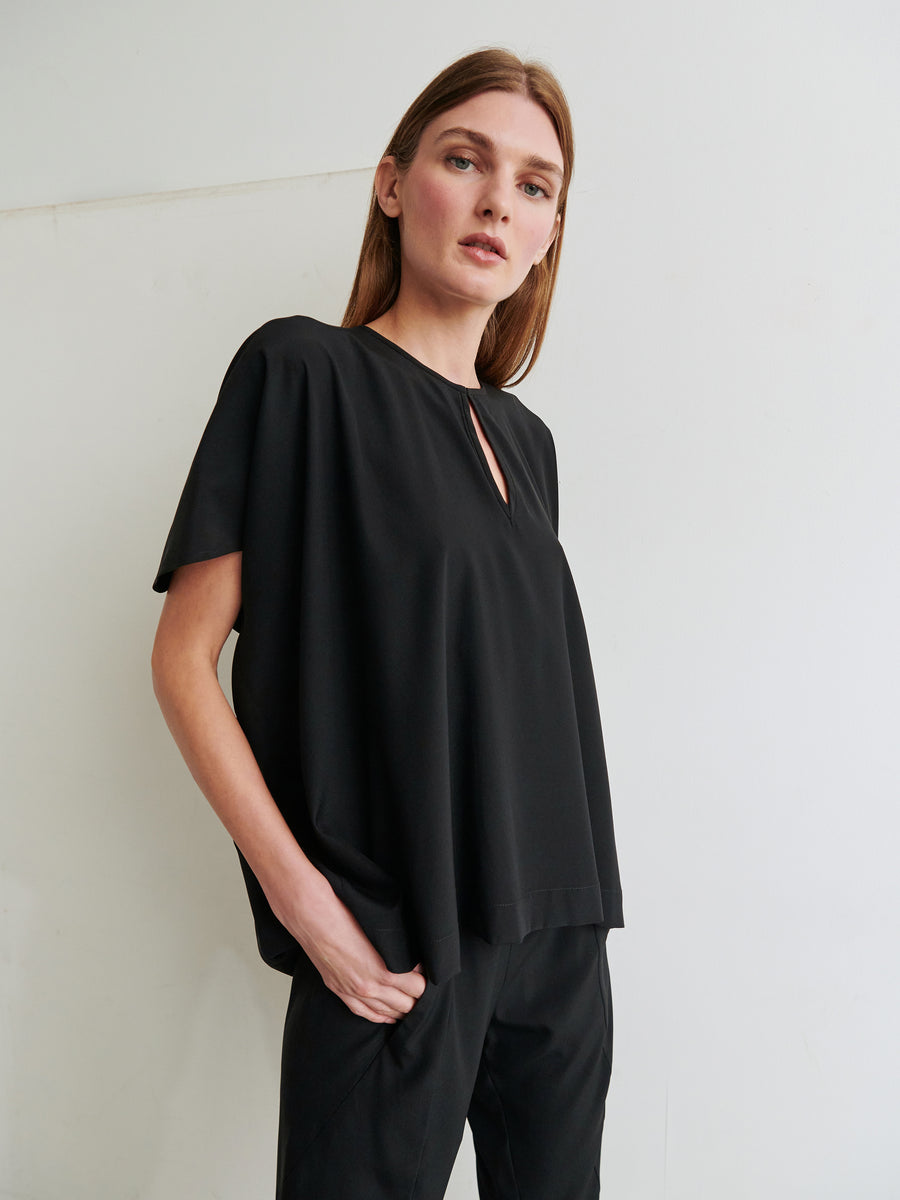 A person with long hair, wearing a loose-fitting Rae Poncho Top in black stretch silk by Zero + Maria Cornejo and black pants, stands with one hand in their pocket. The background is plain white, providing a clean and simple setting. The person gazes slightly upwards with a neutral expression.