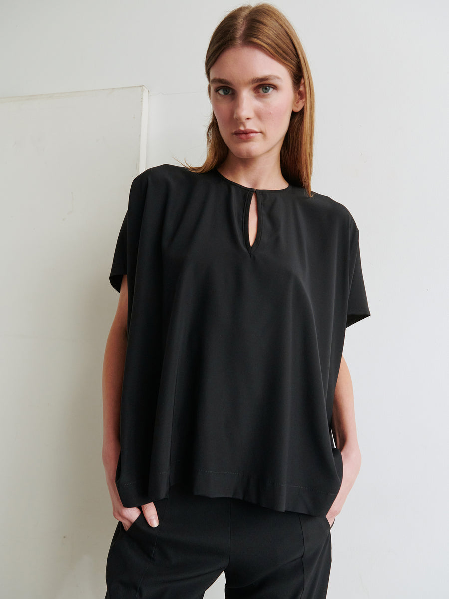 A woman with straight brown hair, wearing the Rae Poncho Top by Zero + Maria Cornejo and matching pants, stands against a white wall. She has her hands in her pockets and looks directly at the camera.