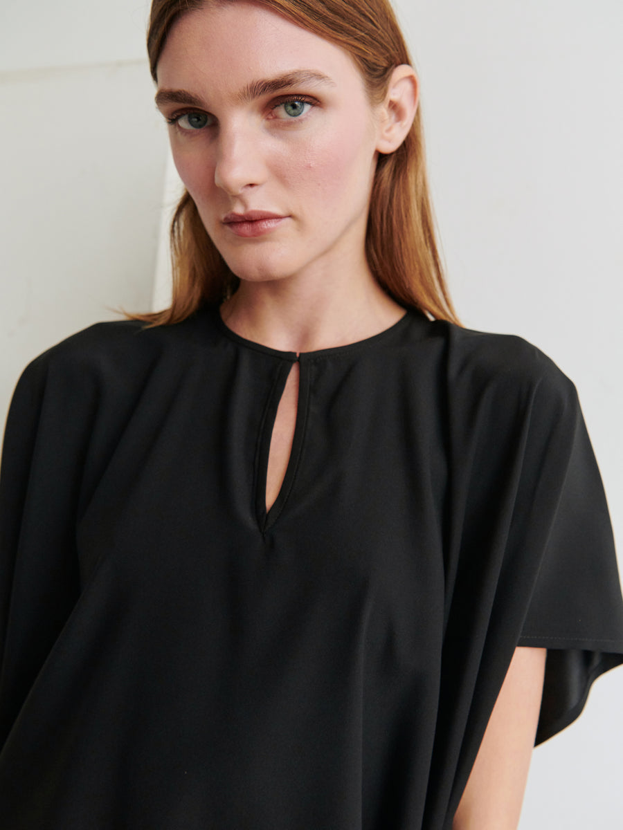 A woman with straight, light brown hair, wearing the black Rae Poncho Top by Zero + Maria Cornejo, featuring a small keyhole neckline, looks directly at the camera. The background is a simple, light-colored wall.