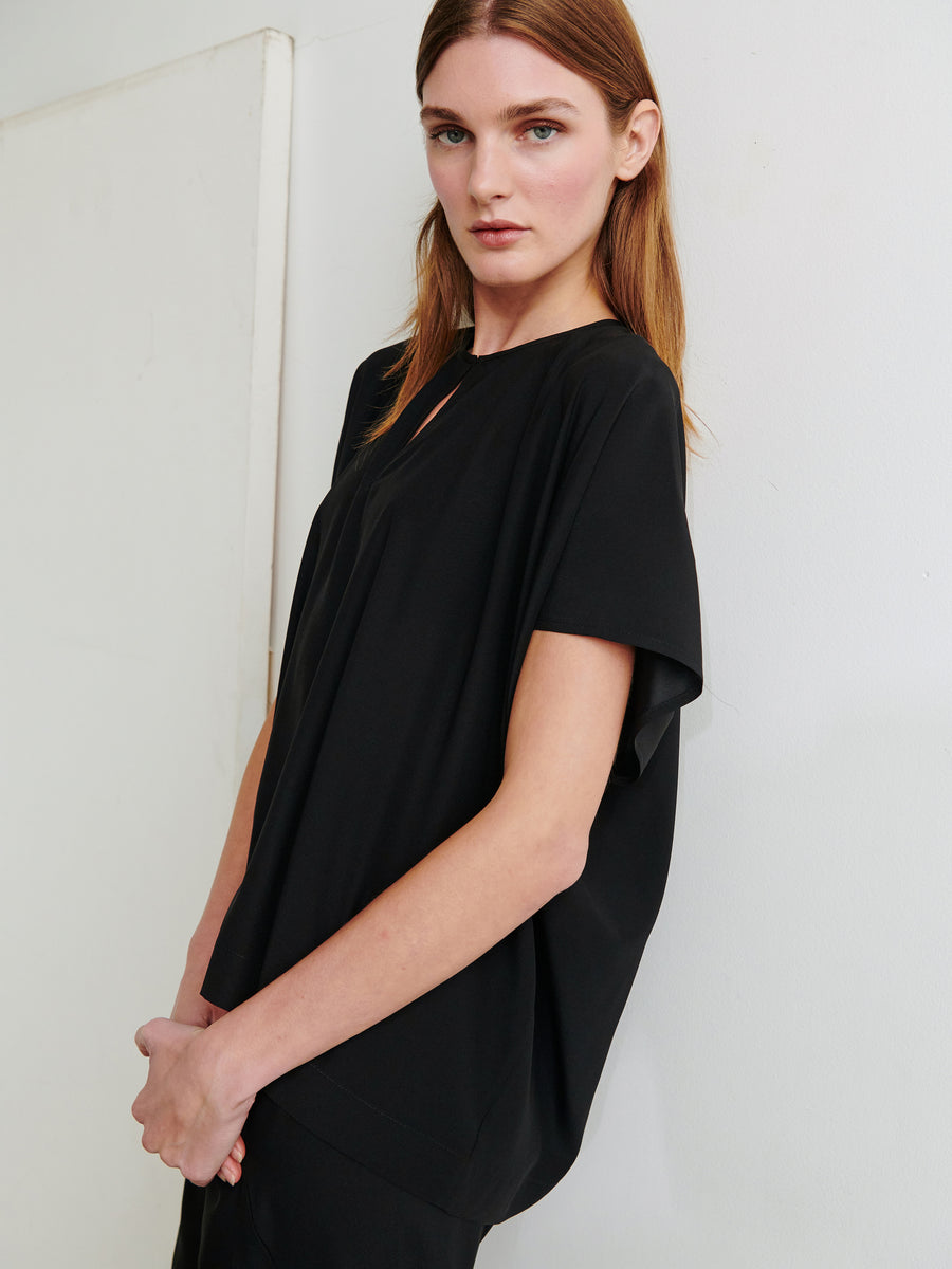 A woman with long, straight, light brown hair is posing against a white wall. She is wearing the Rae Poncho Top by Zero + Maria Cornejo, a loose-fitting black stretch silk top with short sleeves. Her arms are gently crossed and she is looking slightly past the camera, with a calm expression on her face.