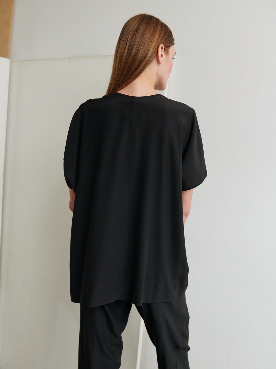 A person with long, straight hair stands facing away, wearing the Rae Poncho Top by Zero + Maria Cornejo and matching black pants. The background is a neutral white wall with a wooden detail on the left.