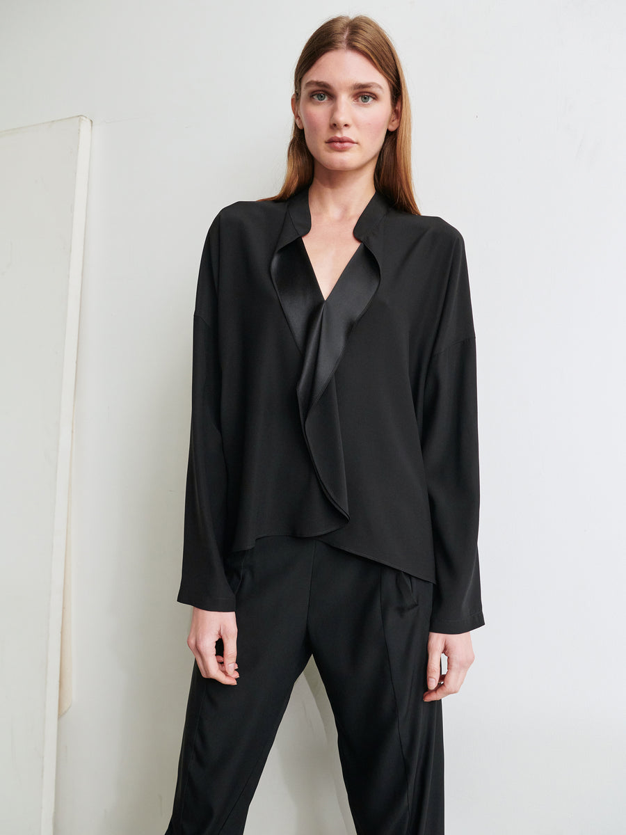 A person with long brown hair is wearing a black, loose-fitting Long-Sleeved Fin Gaban Shirt by Zero + Maria Cornejo. The attire features a low V-neck blouse with draped detailing, paired with matching black trousers. They are standing against a plain, light-colored background and looking directly at the camera.