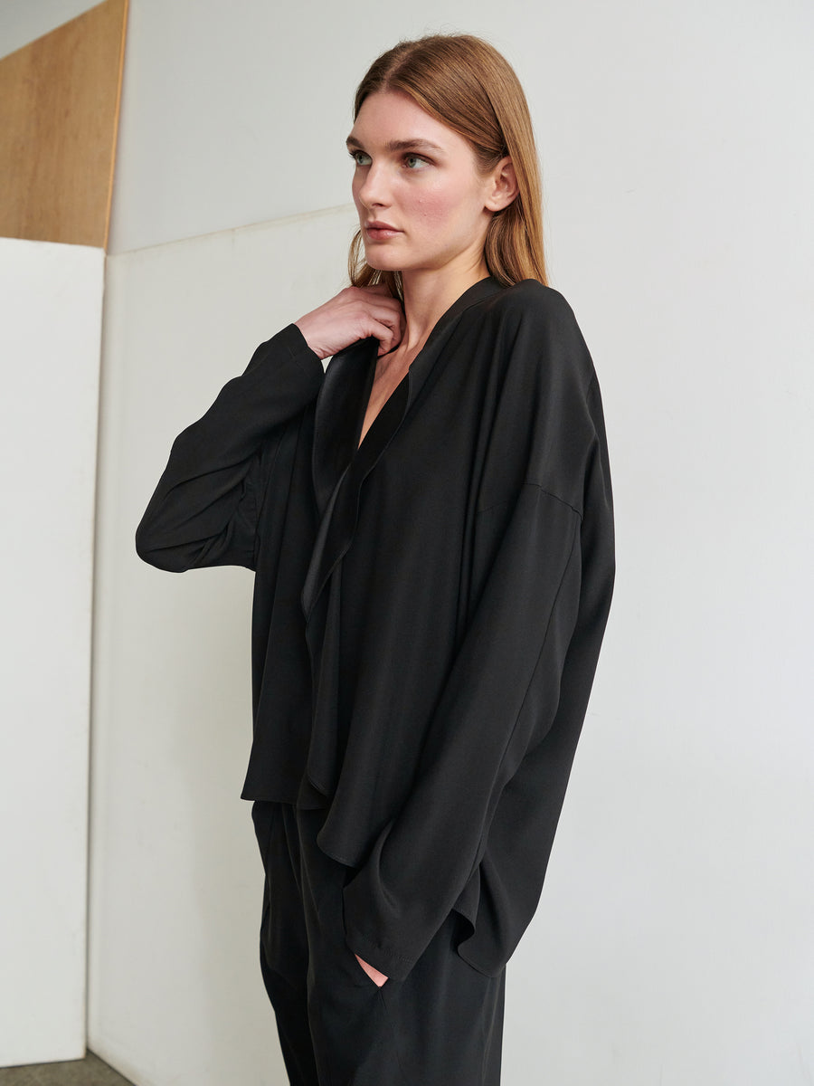 A person with long brown hair gazes to the side, wearing a loose-fitting Long-Sleeved Fin Gaban Shirt from Zero + Maria Cornejo. The background features a white wall and a wooden panel. Their left hand is slightly raised, touching the back of their neck. The overall mood is contemplative.