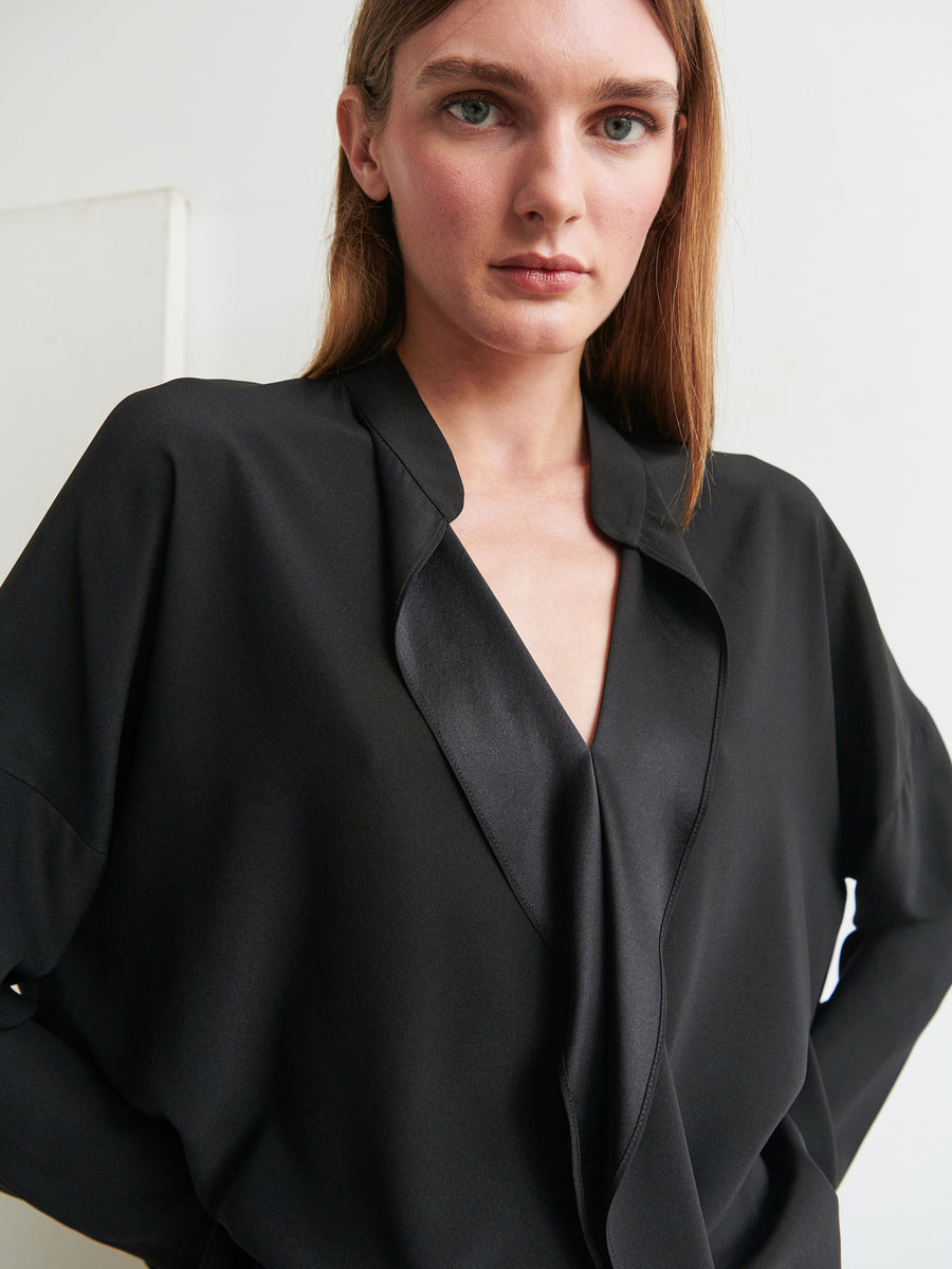 A woman with straight, light brown hair is wearing a black Long-Sleeved Fin Gaban Shirt by Zero + Maria Cornejo. She faces the camera with a neutral expression. The plain white background creates a stark contrast with her dark attire.