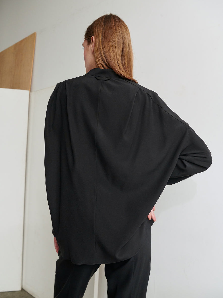 A person with long brown hair, wearing a loose black blazer and pants over a Long-Sleeved Fin Gaban Shirt by Zero + Maria Cornejo, stands with their back to the camera. They have one hand on their hip and are in front of a plain, white wall with a wooden panel to the side.