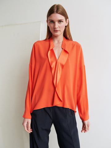 A person with long brown hair is wearing an orange, long-sleeved Fin Gaban Shirt by Zero + Maria Cornejo, featuring a stretch silk charmeuse fabric and a deep V-neck, paired with black pants. The person is standing in front of a plain white wall.