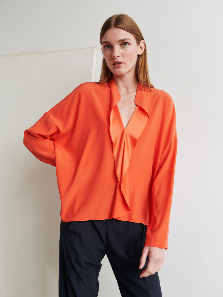 A person with long brown hair stands in front of a plain white wall. They wear the loose-fitting, bright orange Long-Sleeved Fin Gaban Shirt by Zero + Maria Cornejo paired with black trousers. One hand rests on their hip as they look slightly to the side.