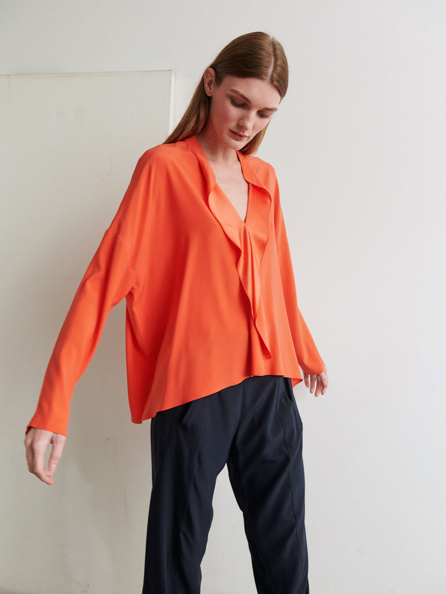 A person with long brown hair wearing an oversized, long-sleeved Fin Gaban Shirt by Zero + Maria Cornejo and black pants stands against a plain white background. They have their head tilted to one side and arms slightly extended, creating a relaxed and poised stance.