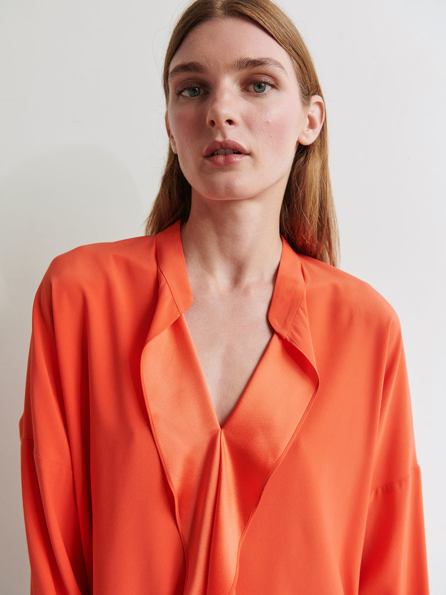 A person with long, light brown hair stands against a plain background. They are wearing an orange Long-Sleeved Fin Gaban Shirt by Zero + Maria Cornejo, featuring a deep V-neckline. The individual gazes into the camera with a neutral expression.