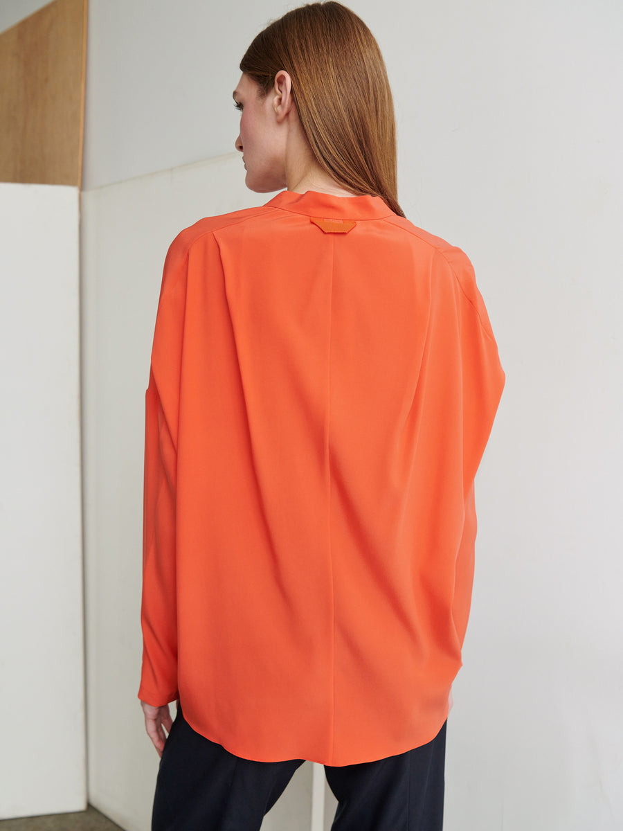 A person with long brown hair is seen from the back, wearing a loose-fitting Long-Sleeved Fin Gaban Shirt in orange by Zero + Maria Cornejo. They are standing in a room with a gray wall and a wooden panel to the left, also dressed in black pants.