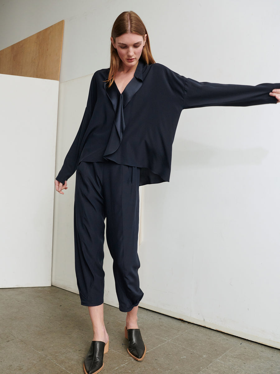 A person with long straight hair is standing indoors against a white and wooden-paneled wall. They are wearing a loose-fitting, navy blue outfit featuring the Long-Sleeved Fin Gaban Shirt by Zero + Maria Cornejo and cropped pants. With one arm outstretched, they are also sporting black slip-on shoes.