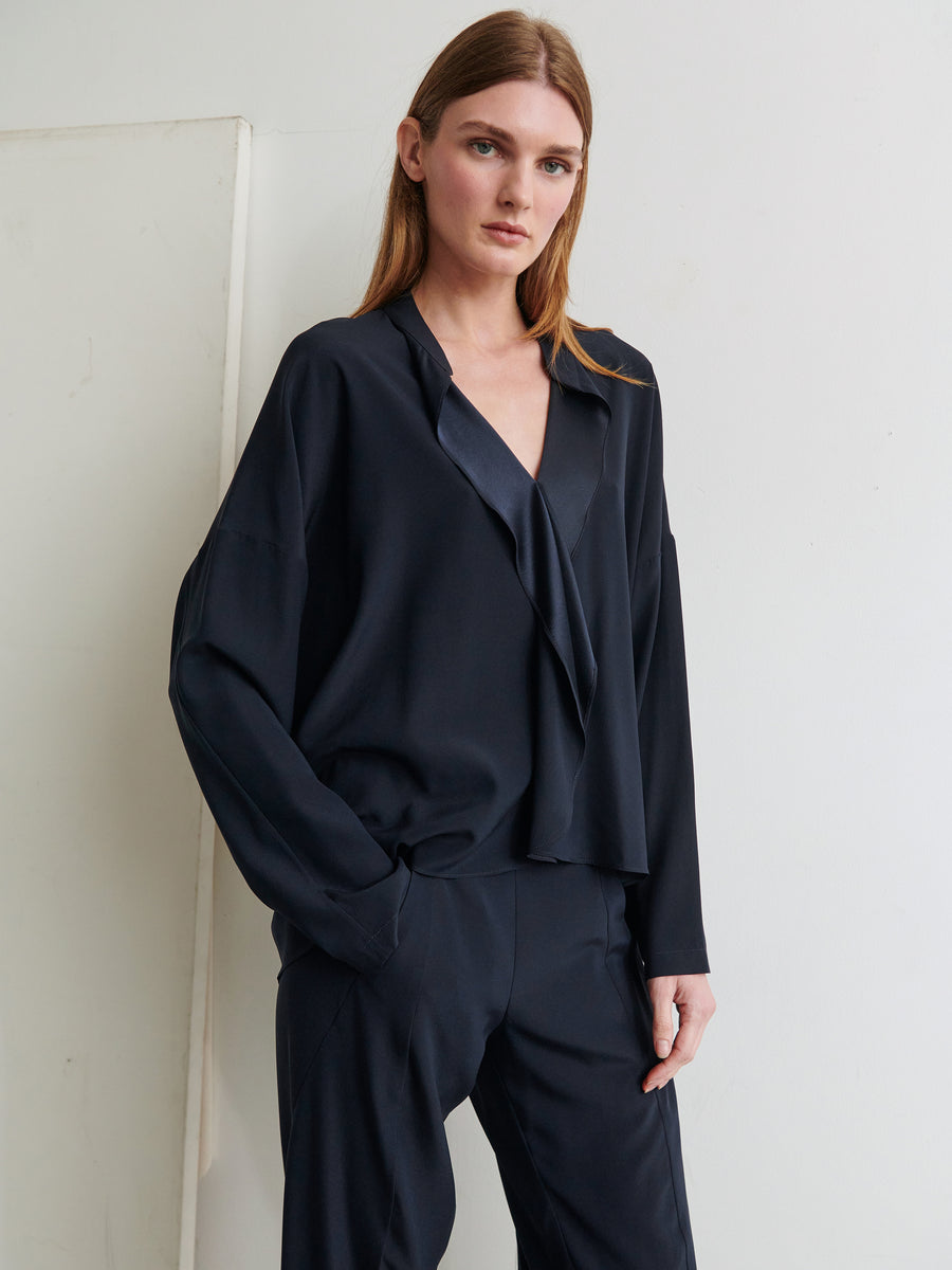 A woman with long blonde hair stands against a white wall, wearing a loose-fitting, dark navy Long-Sleeved Fin Gaban Shirt from Zero + Maria Cornejo and matching trousers. The blouse features draped ruffle details, and she has a neutral expression with one hand resting in her pocket.