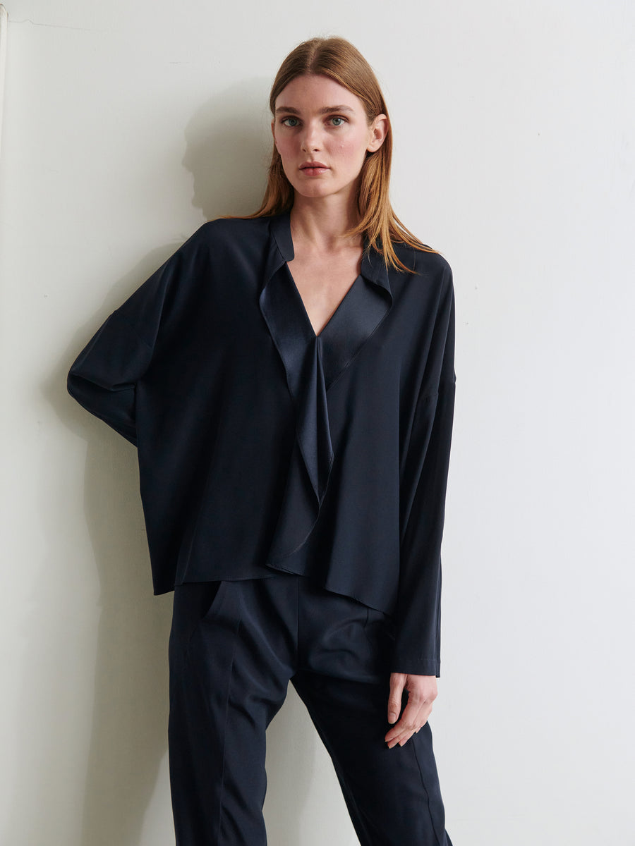 A person with long, straight hair is standing against a light wall. They are wearing a loose-fitting, dark Long-Sleeved Fin Gaban Shirt with a deep V-neck and matching dark pants from Zero + Maria Cornejo. Their left arm rests on the wall, and they have a neutral expression on their face, paired with the elegance of their stretch silk charmeuse shirt.