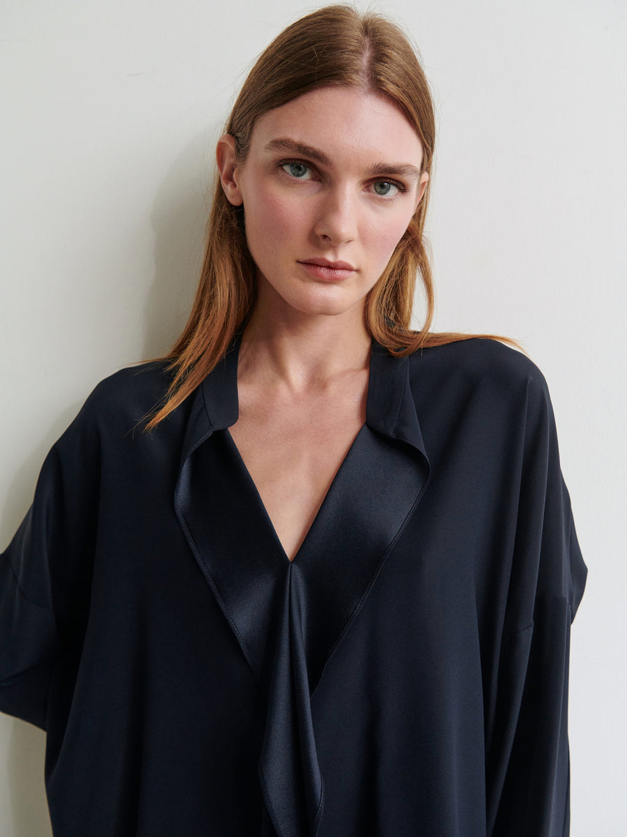 A person with straight, light brown hair is standing against a plain white wall. They have a neutral expression and are wearing the Long-Sleeved Fin Gaban Shirt in a dark hue from Zero + Maria Cornejo, featuring a loose fit, stretch silk charmeuse fabric, deep V-neck, and collar.