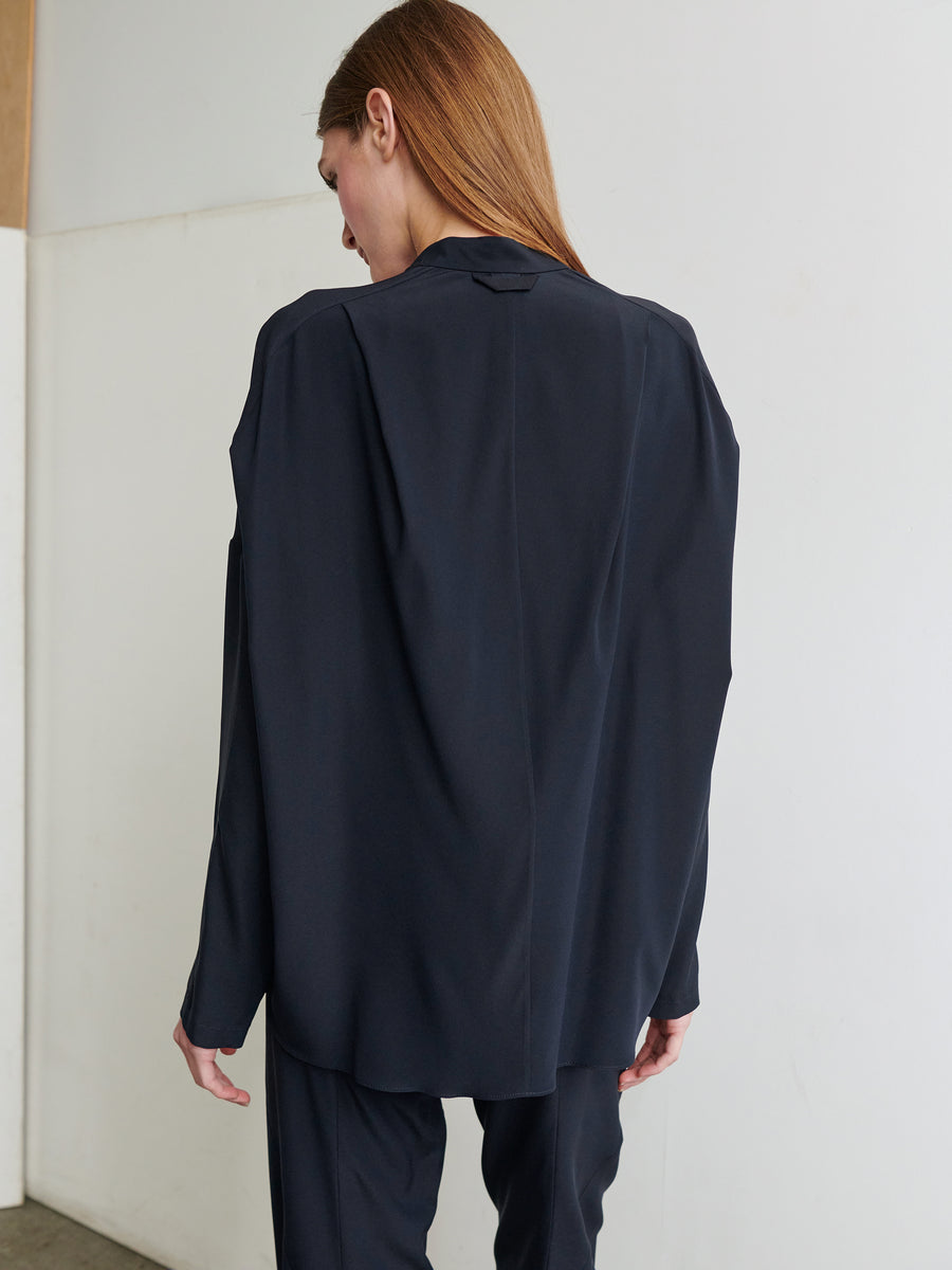 A person with long, straight hair is standing with their back to the camera, wearing a loose-fitting, dark-colored Long-Sleeved Fin Gaban Shirt from Zero + Maria Cornejo. The background is minimalist, featuring light-colored walls and a wooden panel.