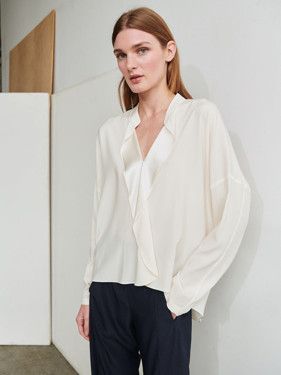 A person with long, light brown hair stands in a minimalist room wearing a white, loosely fitted Long-Sleeved Fin Gaban Shirt by Zero + Maria Cornejo with a V-neck and black pants. They are looking directly at the camera with a neutral expression, one hand resting at their side and the other partially in a pocket.