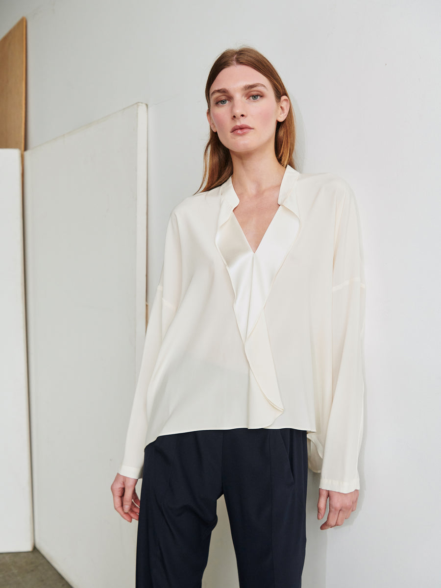 A person with long hair is standing against a white wall looking at the camera. They are wearing the Long-Sleeved Fin Gaban Shirt from Zero + Maria Cornejo, featuring a loose fit and draped ruffle detail, paired with black trousers. The setting appears to be minimalistic and indoors.