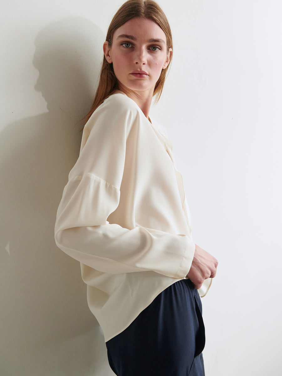 A person with long hair is standing against a white wall, gazing into the distance. They are wearing a Long-Sleeved Fin Gaban Shirt in cream-colored stretch silk charmeuse by Zero + Maria Cornejo alongside dark pants. The overall tone of the image is minimalistic and serene.