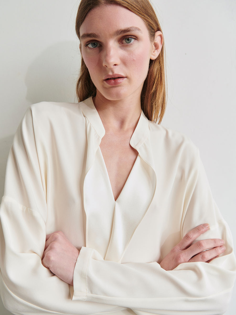 A woman with straight, light brown hair is wearing a Long-Sleeved Fin Gaban Shirt from Zero + Maria Cornejo, featuring a draped ruffle detail in cream-colored stretch silk charmeuse. She is standing against a white background, looking directly at the camera with her arms crossed.