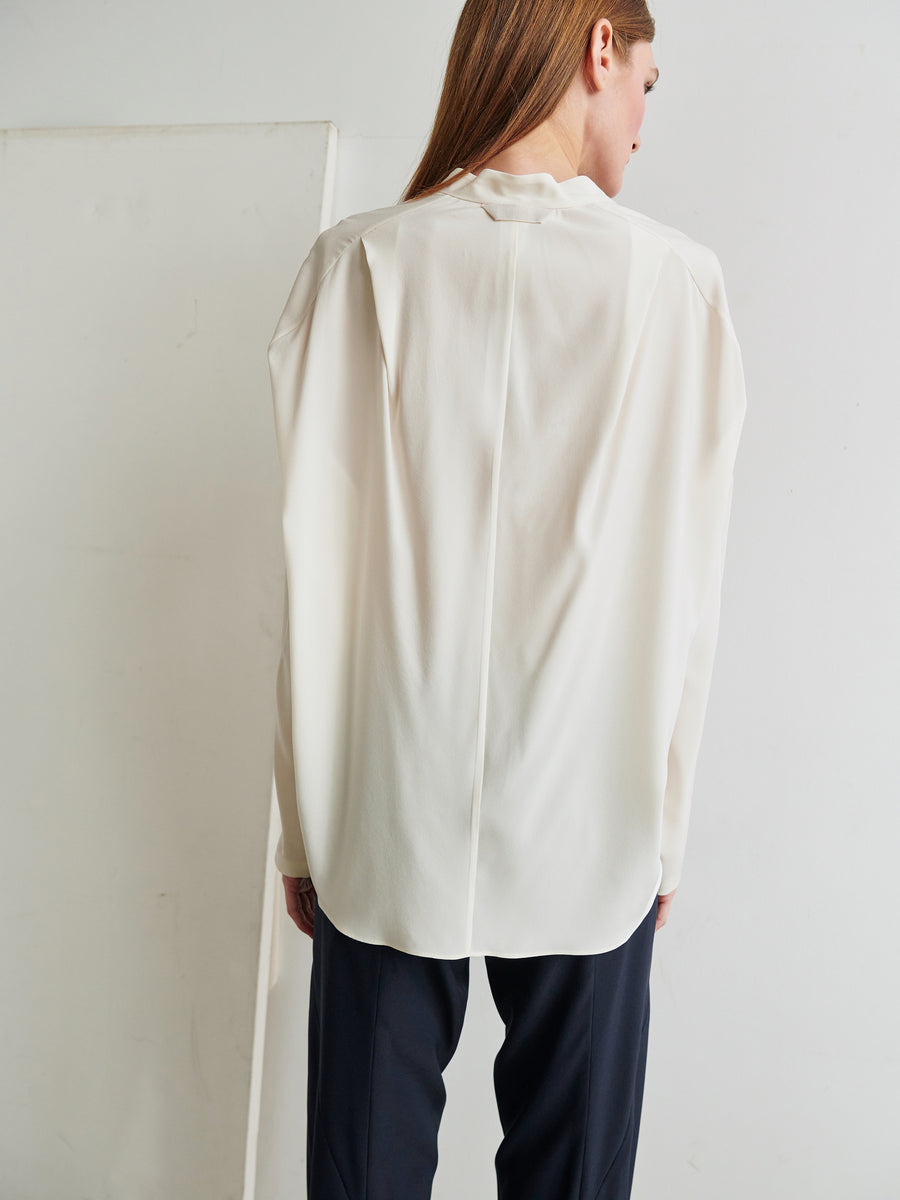 A person with long hair is seen from the back, wearing a white, loose-fitting Long-Sleeved Fin Gaban Shirt by Zero + Maria Cornejo and dark pants. The setting appears to be minimalist with plain white walls and a neutral background.