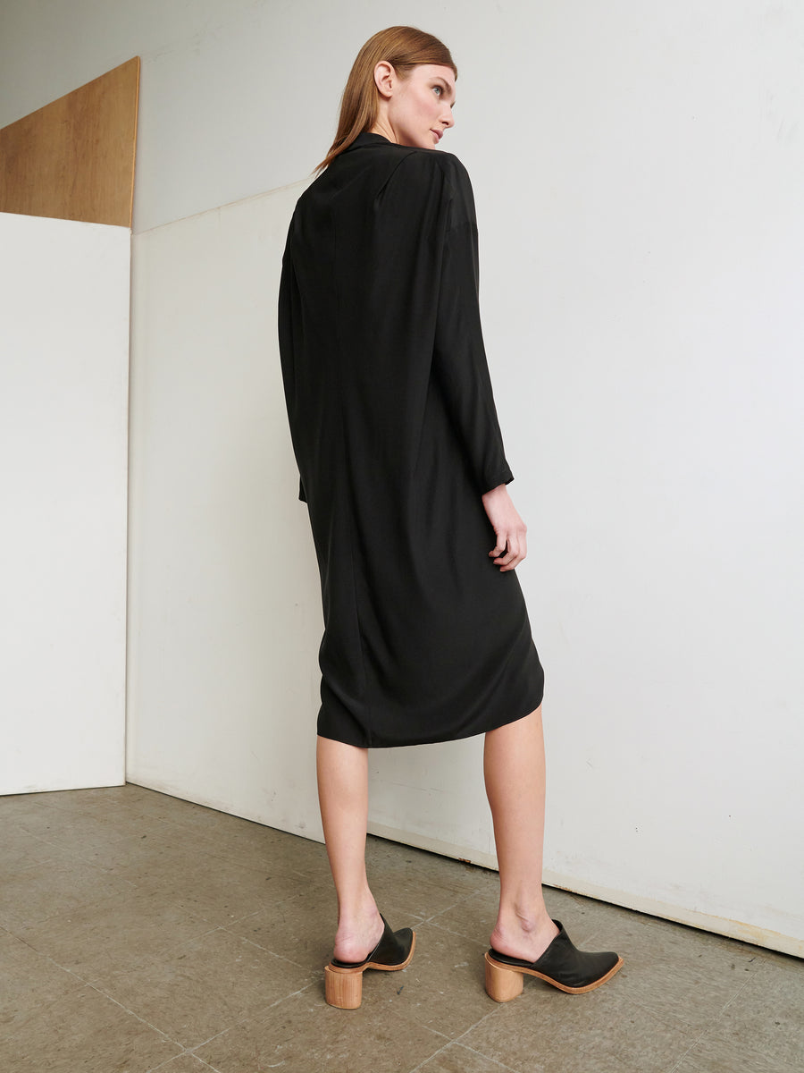 A person with long blonde hair is shown standing, facing away from the camera, wearing a Long-Sleeved Fin Gaban Dress from Zero + Maria Cornejo that falls to mid-calf and black heeled mules with wooden soles. The background features white and wooden partitions and a concrete floor.