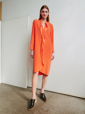 A person with long hair stands indoors wearing an orange Long-Sleeved Fin Gaban Dress by Zero + Maria Cornejo, paired with black slip-on shoes featuring a low block heel. The background showcases a plain white wall and a section of wooden flooring.