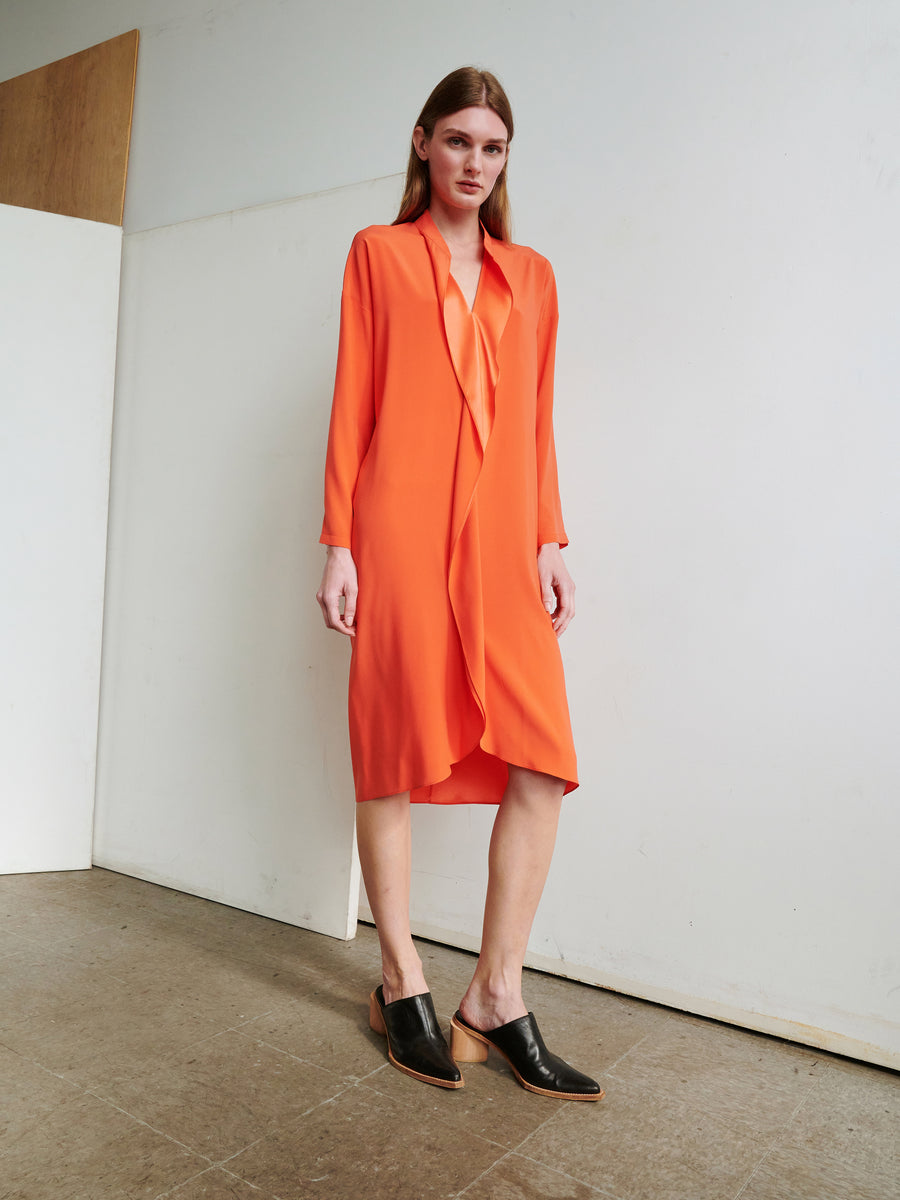 A person with long hair stands against a plain wall, wearing the vibrant orange, knee-length Long-Sleeved Fin Gaban Dress by Zero + Maria Cornejo. The dress features a draped front and subtle ruffle details that add elegance. They pair it with black slip-on shoes with a low wooden heel. The floor beneath them is tiled.