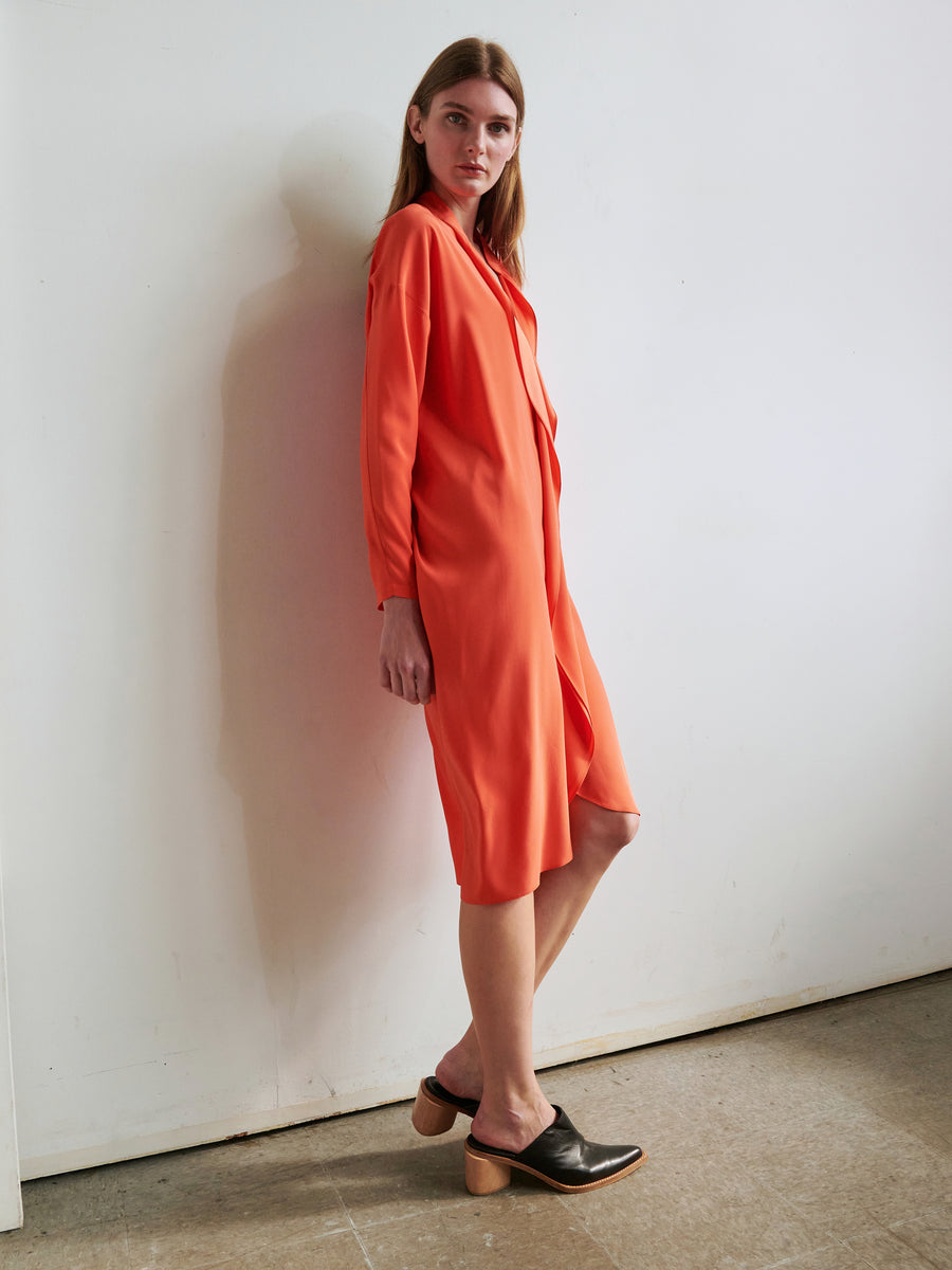 A person with long hair stands against a wall, wearing an orange Long-Sleeved Fin Gaban Dress by Zero + Maria Cornejo, paired with black shoes featuring wooden heels. The setting appears minimalistic with a plain background and concrete floor.