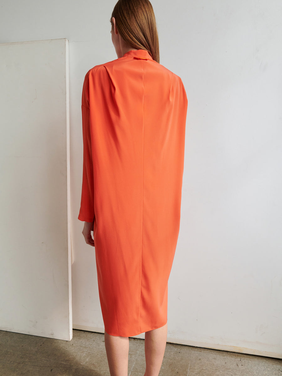 A person with long light brown hair and fair skin is standing with their back to the camera, wearing a knee-length Long-Sleeved Fin Gaban Dress in vibrant orange from Zero + Maria Cornejo. The background is minimalistic, featuring a plain white wall and a white door or panel on the left side.