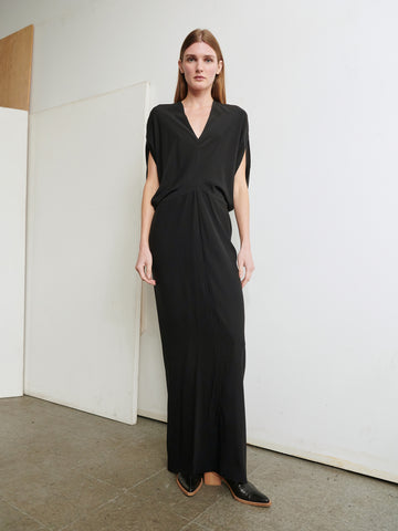 A person with long hair stands against a white wall wearing the sleek, slim fit Long Reni Dress by Zero + Maria Cornejo, featuring a V-neck and short sleeves. They have black pointed shoes, and the background includes a segment of a wooden panel and a gray tiled floor.