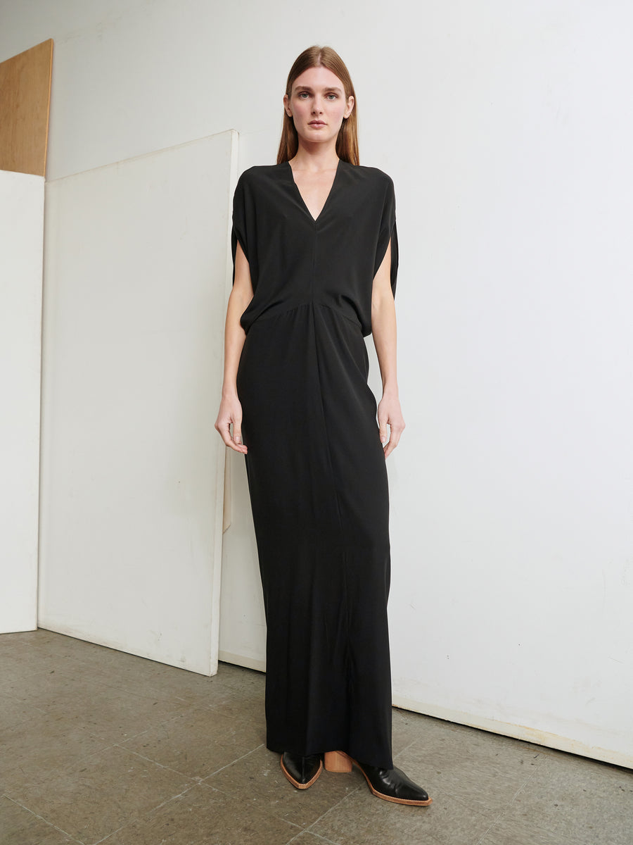 A person with long hair stands against a white wall wearing the sleek, slim fit Long Reni Dress by Zero + Maria Cornejo, featuring a V-neck and short sleeves. They have black pointed shoes, and the background includes a segment of a wooden panel and a gray tiled floor.