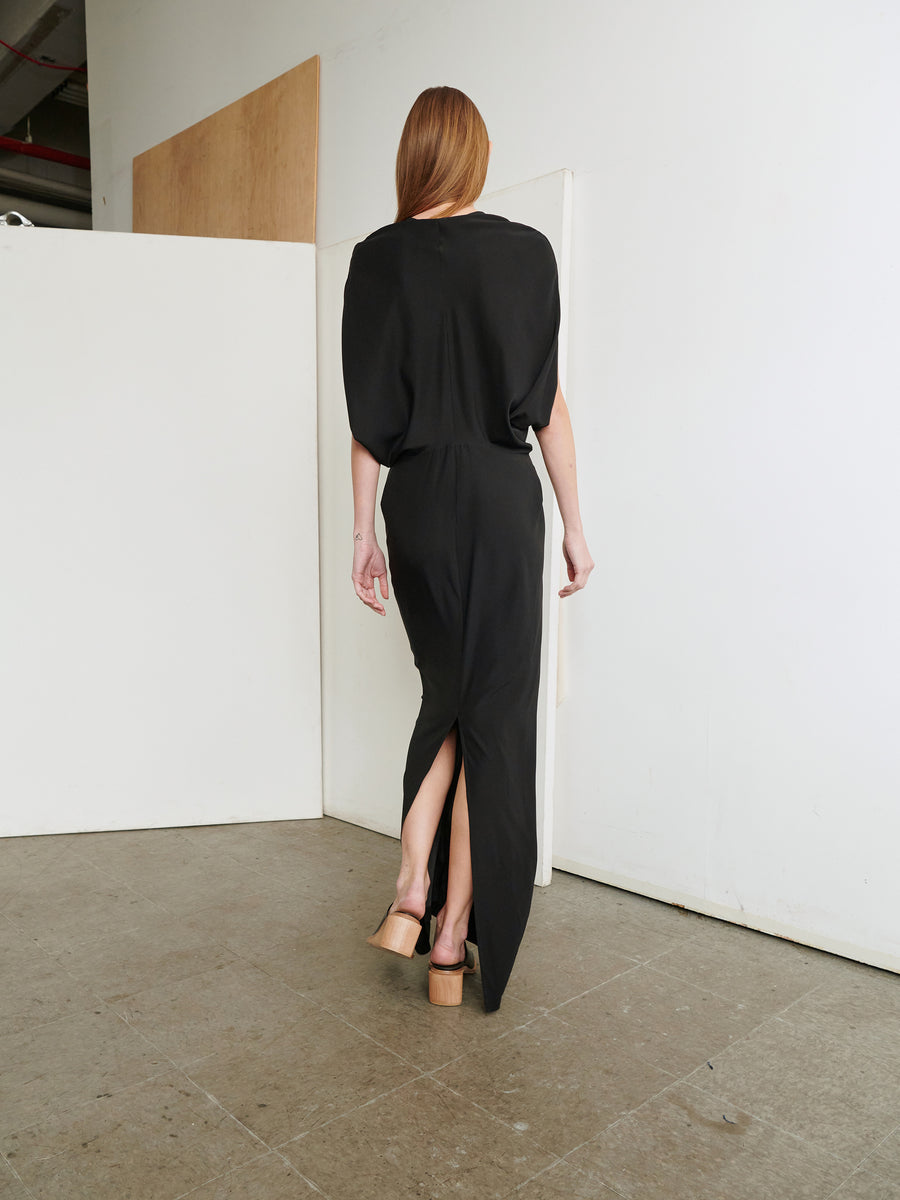 A person with long auburn hair is seen from behind, showcasing a slim fit dress, styled with nude high heels. The outfit—a Long Reni Dress by Zero + Maria Cornejo, featuring a slit at the back—contrasts elegantly with the minimal background of white walls and a wooden panel.