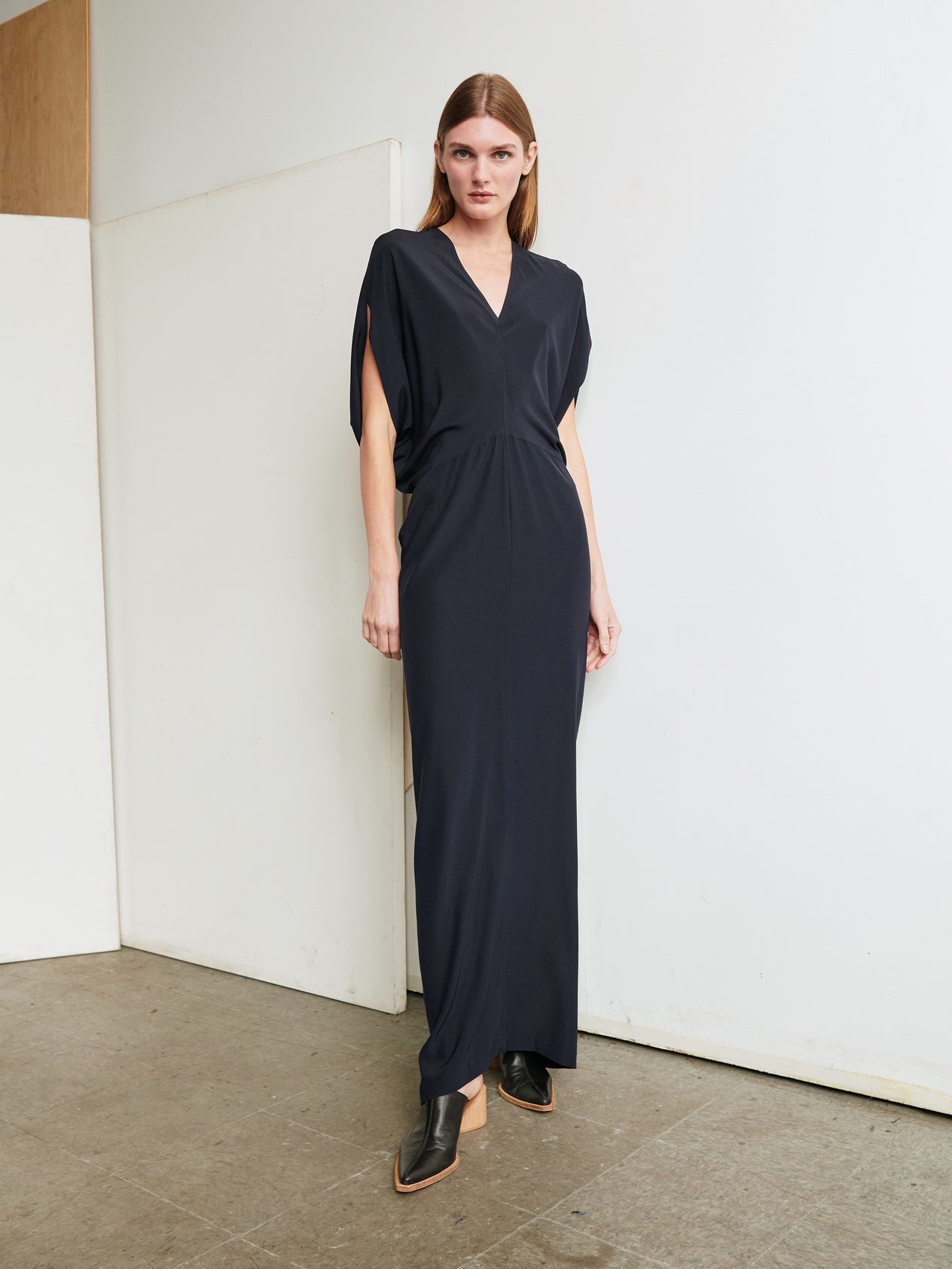 A person with long hair stands against a white wall wearing the sleek, slim fit Long Reni Dress by Zero + Maria Cornejo, featuring a V-neck and short sleeves. They have black pointed shoes, and the background includes a segment of a wooden panel and a gray tiled floor.