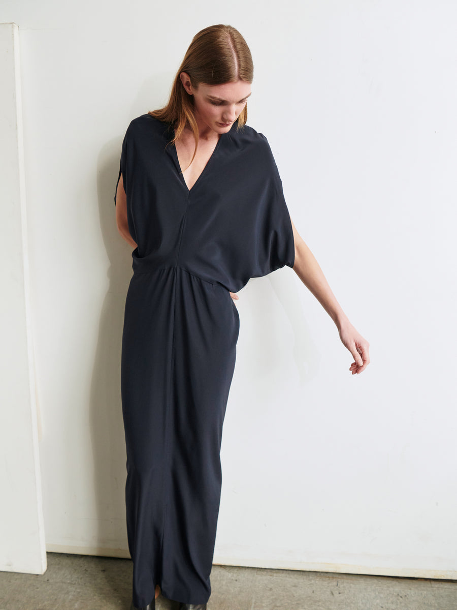 A person with long hair is wearing the elegant Long Reni Dress from Zero + Maria Cornejo, featuring a V-neck and short, flowing sleeves. The person is slightly leaning to one side against a white wall, looking down with one arm hanging by their side and the other touching the wall.