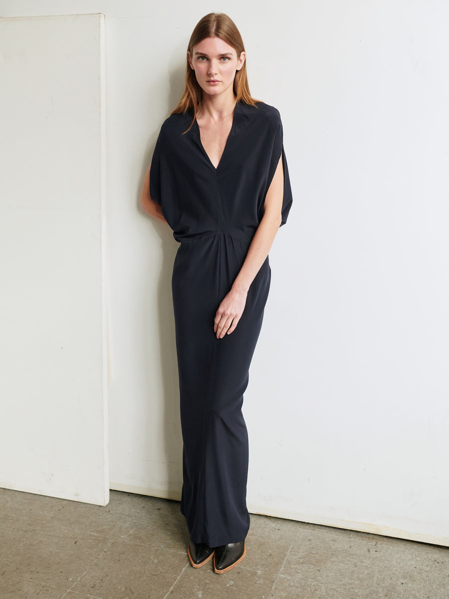 A woman with straight hair stands against a white wall on an unfinished floor. She is wearing the Long Reni Dress by Zero + Maria Cornejo, along with pointed-toe shoes. Her hands are relaxed by her sides, and she looks directly at the camera with a neutral expression.