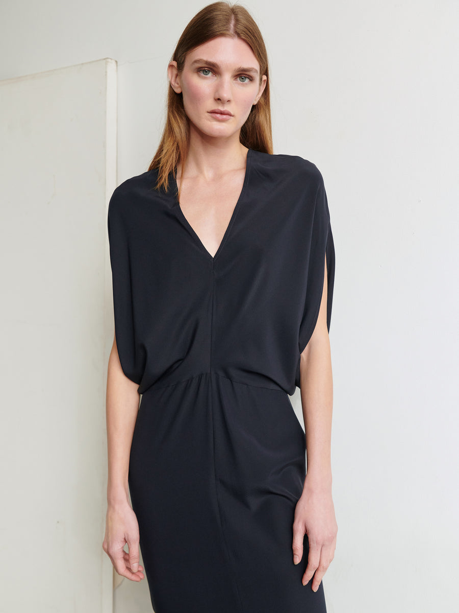 A person with long, straight hair is wearing the Long Reni Dress from Zero + Maria Cornejo, a simple black stretch silk charmeuse dress featuring a deep V-neck and short sleeves. They are standing against a plain, light-colored wall and looking directly at the camera with a neutral expression.