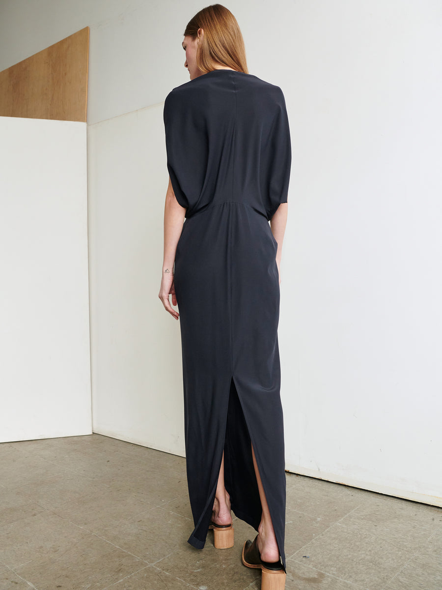 A person with long, light brown hair is standing indoors, wearing a Zero + Maria Cornejo Long Reni Dress made from dark-colored stretch silk charmeuse with short sleeves. The individual is viewed from behind in a minimalist room with a white and wooden interior, showcasing the elegant back slit of the slim skirt.