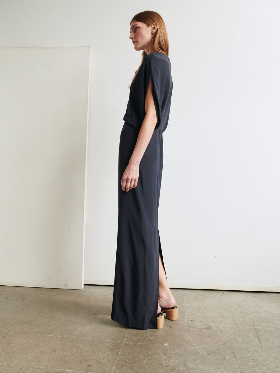 A person with long hair is seen standing in profile view, adorned in a dark-colored Long Reni Dress by Zero + Maria Cornejo. The stretch silk charmeuse dress highlights a slim skirt with a back slit, complemented by heeled shoes. The minimalist background features a plain white wall and a subtly textured grey floor.