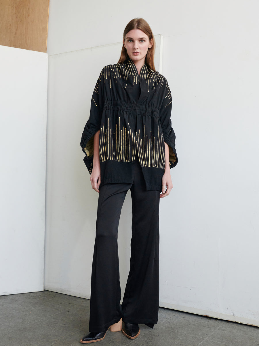 A woman with long hair stands indoors by a white wall and wooden panel. She is dressed in the Evia Shrug by Zero + Maria Cornejo, a black outfit featuring a long top adorned with gold vertical lines resembling a metallic sparkler design, paired with black flared pants. She looks directly at the camera with a neutral expression, her hands resting by her sides.