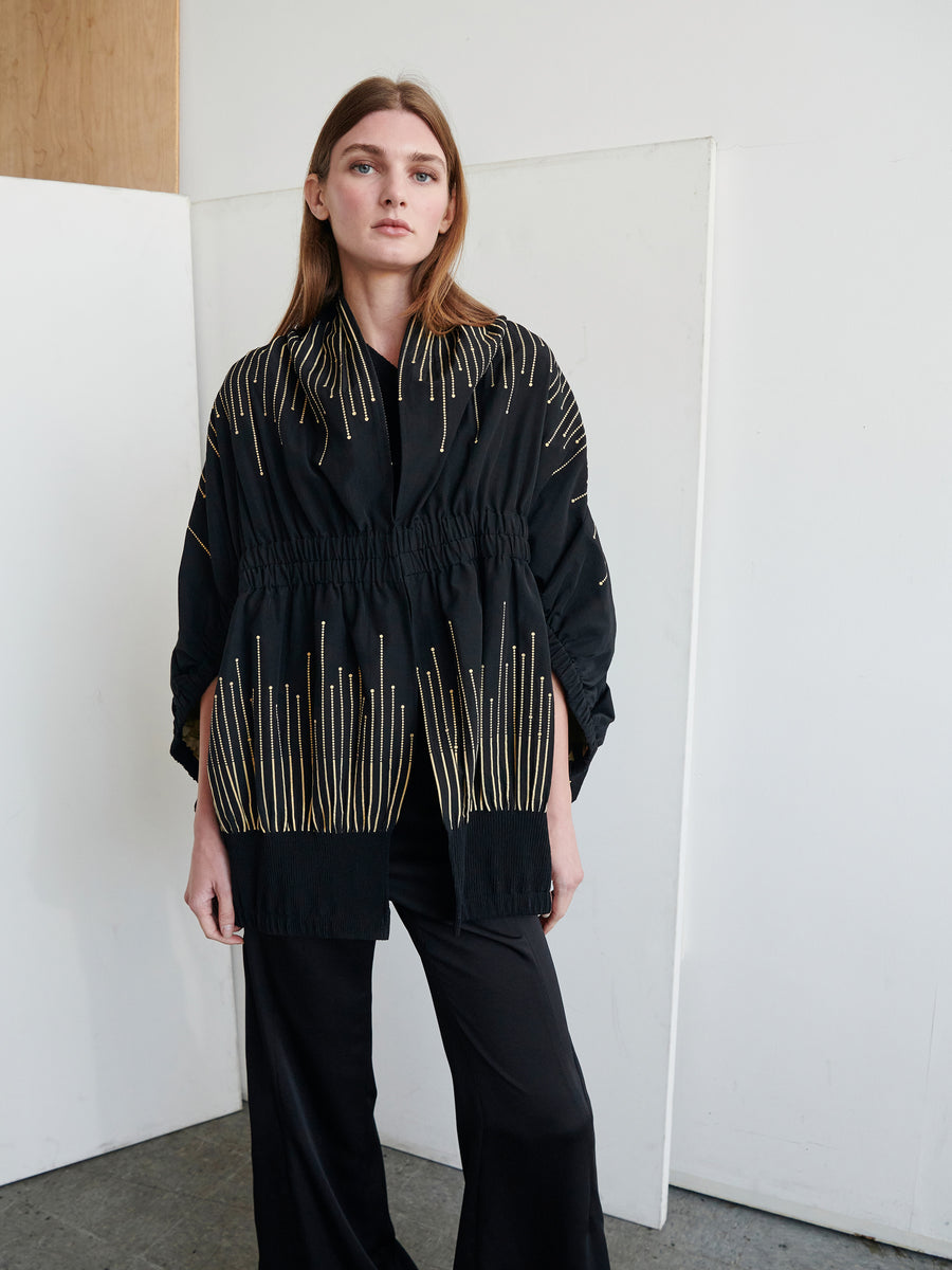 A person with long brown hair stands indoors against a minimalist background. They are wearing the Evia Shrug by Zero + Maria Cornejo, a black kimono-style top adorned with gold vertical stripes reminiscent of a metallic sparkler design, paired with black pants. The person gazes directly at the camera with a neutral expression.