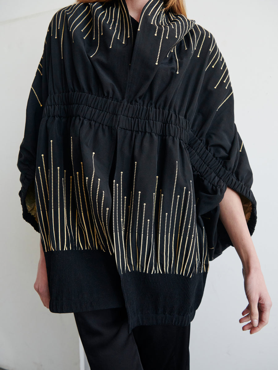 A person wearing the Evia Shrug from Zero + Maria Cornejo, a black kimono-style garment with intricate vertical golden embroidery, featuring a ruched waistline and loose, three-quarter-length sleeves. The recycled fibers give the outfit an eco-friendly touch. The person stands against a plain white background.