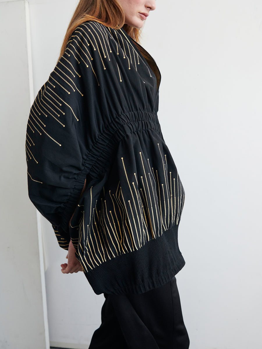 A person wearing the Evia Shrug by Zero + Maria Cornejo, a black, loose-fitting garment featuring thin gold vertical lines in a metallic sparkler design. The shrug has an elasticated waist and voluminous sleeves. The background is plain white, and the person's face is partially out of frame.