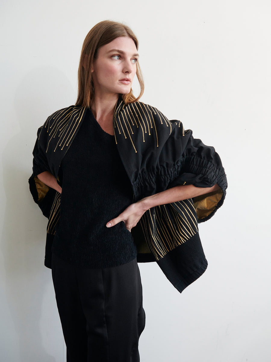 A woman with long brown hair stands against a light-colored wall. She is dressed in the Evia Shrug from Zero + Maria Cornejo, an elegant black outfit made of recycled polyester, featuring intricate gold embroidery on the shoulders and sleeves. She gazes contemplatively into the distance with her hands resting on her hips.